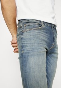 Vaqueros de mezclilla desgastados en azul claro con un leve desvanecimiento, diseño de cinco bolsillos y costuras amarillas a lo largo de las costuras y los bolsillos.