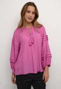 Blusa rosa con maniche lunghe, caratterizzata da accentuazioni arricciate, un collo tirato e dettagli con frange, realizzata in un tessuto leggero.
