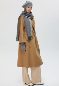 Manteau en laine beige avec une taille ceinturée, associé à un pantalon large beige clair. Les accessoires comprennent une écharpe grise et un béret assorti.