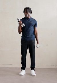 Black athletic pants and a navy blue short-sleeve shirt, holding a silver dumbbell and a lighter weight, standing on a light gray floor.