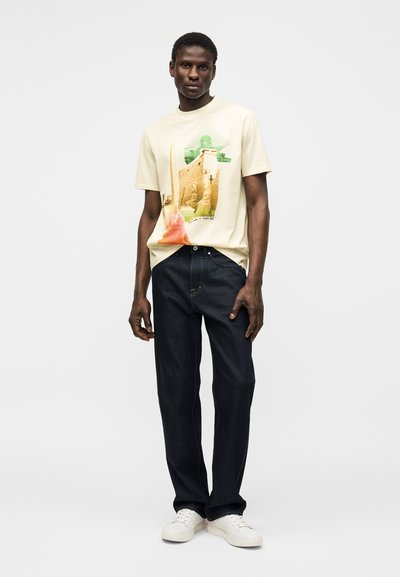 Άνδρας με κρέμ γραφικό μπλουζάκι με αιγυπτιακό σχέδιο, σκούρο τζιν και λευκά αθλητικά παπούτσια, στέκεται μπροστά σε απλό φωτεινό φόντο.