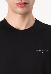 Primo piano di una persona che indossa una maglietta nera con scollo a girocollo con il logo "TOMMY JEANS" e un piccolo emblema a bandiera sul petto.