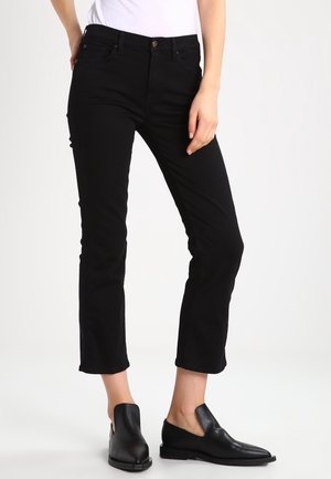 Mujer con pantalones negros ajustados y recortados y zapatos negros sin cordones, de pie con una mano apoyada en su muslo.