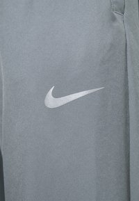 Nahaufnahme eines grauen Stoffes mit einem reflektierenden weißen Nike-Swoosh-Logo, das darauf gedruckt ist und einen Teil der Naht einer Kleidungsstücktasche zeigt.