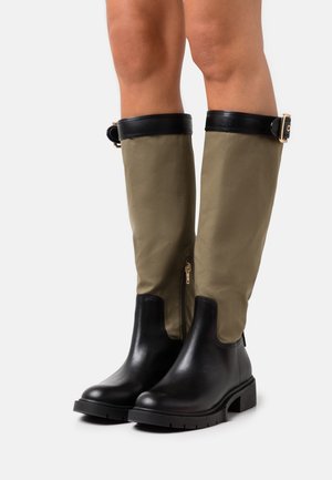Botas tobilleras de cuero negro con una parte superior de tela caqui, que cuentan con un cierre lateral y una correa negra con una hebilla dorada en la parte superior.