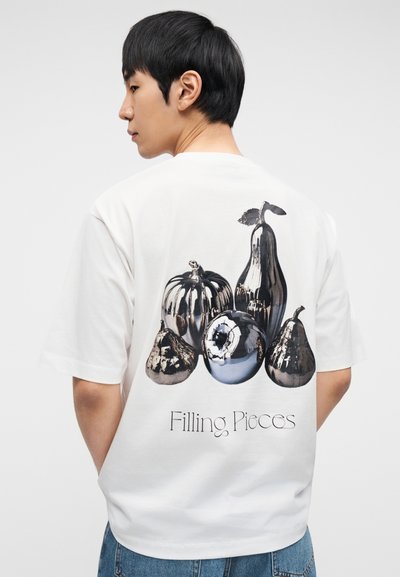 Λευκό βαμβακερό μπλουζάκι με εκτύπωση μεταλλικών φρούτων στην πλάτη. Το σχέδιο περιλαμβάνει αχλάδι, κολοκύθα και μήλο. Εμφανίζεται το λογότυπο της μάρκας.