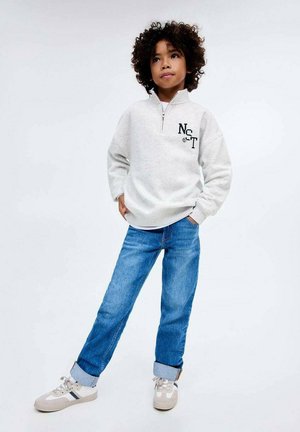 Pullover grigio in pile con zip a metà, logo "NCT" ricamato, abbinato a jeans in denim blu con risvolto e scarpe da ginnastica grigie con dettagli a strisce.