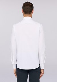 Witte langwerpige blouse met een klassieke kraag, subtiele textuur en afgeronde zoom. Voorzien van knoopsluiting bij de mouwen en een eenvoudig, strak ontwerp.