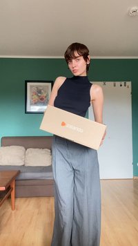 Bruine Zalando-doos vastgehouden voor de camera, met een oranje logo. Een persoon in een zwarte mouwloze top en grijze wijde broek staat in een kamer.