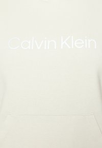 Sudadera con capucha color crema hecha de tela suave, con un bolsillo delantero y un sutil logo blanco de "Calvin Klein" en el pecho.