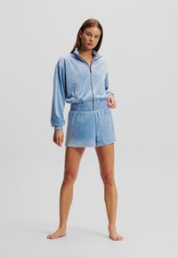 Det blåa velour-träningssetet består av en zip-up jacka med sidofickor och matchande shorts. Tyget verkar mjukt och lätt glänsande.