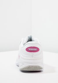 Vit K-Swiss sneakers visade bakifrån med rosa logotypdetalj ovanför en texturerad grå sula på en vit yta.