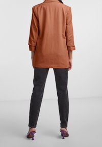 Rostorange blazer med trekvartsärm, kombinerad med mörkgrå slim-fit byxor och lila klacksandaler. Slät tygtextur.