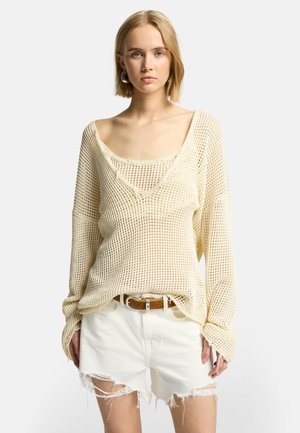 Cremfarbener, locker gestrickter Pullover mit weitem Ausschnitt über einem engen Tank-Top, kombiniert mit zerrissenen weißen Denim-Shorts und einem braunen Gürtel.