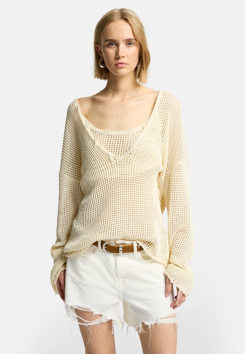 Cremfarbener, locker gestrickter Pullover mit weitem Ausschnitt über einem engen Tank-Top, kombiniert mit zerrissenen weißen Denim-Shorts und einem braunen Gürtel.
