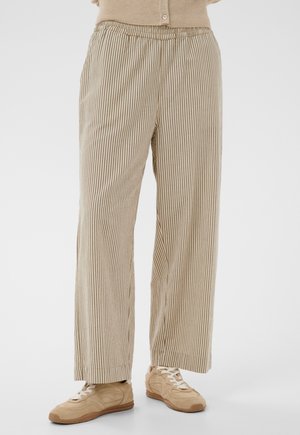 Beige a bílé vertikálně pruhované kalhoty širokých nohavic nošené s béžovými teniskami a béžovým zapnutým topem, pohled od pasu k nohám.