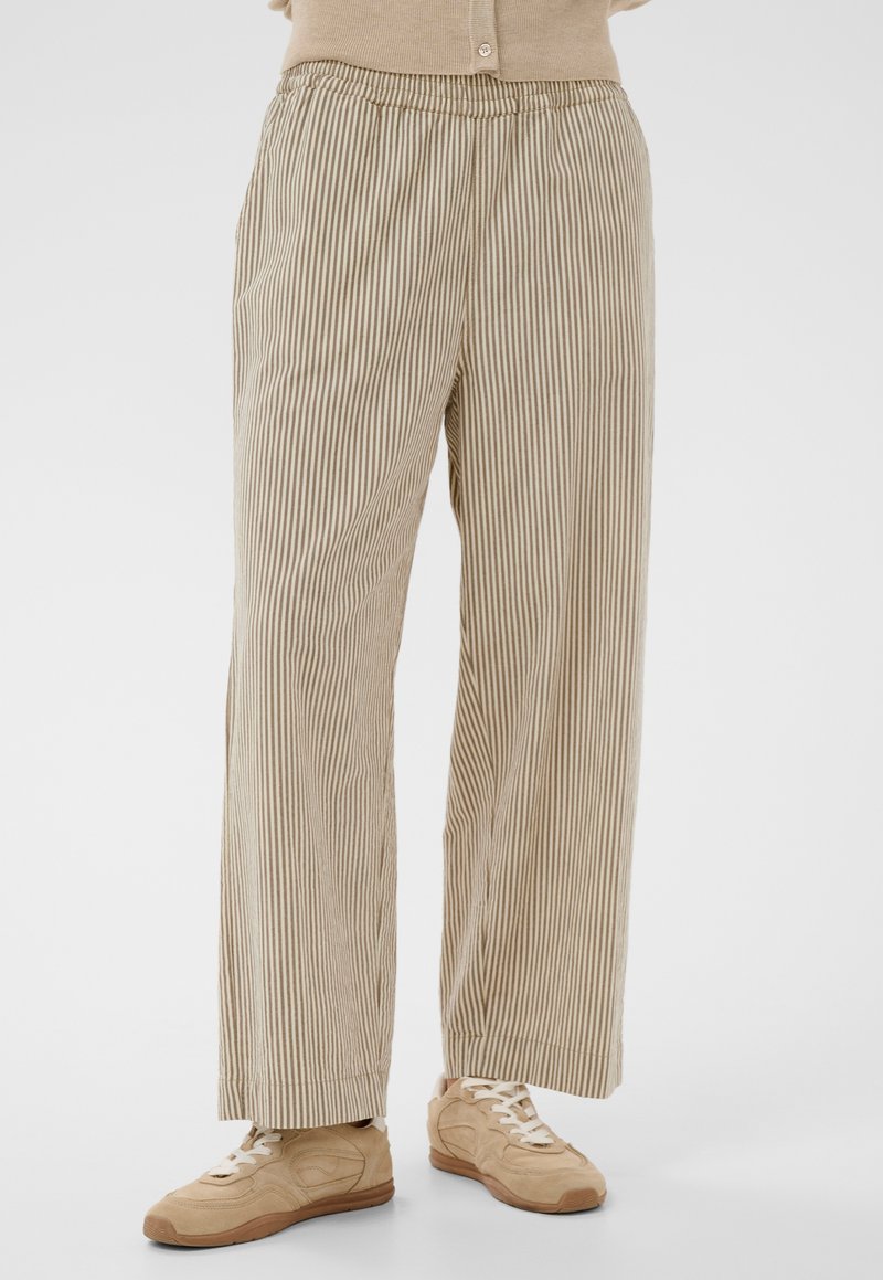Béžové a biele nohavice s širokými nohavicami s vertikálnymi pruhmi, nosené s béžovými teniskami a béžovým zapínaným topom, pohľad od pásu po chodidlá.