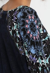 Spalla e parte superiore della schiena di una persona che indossa un indumento scuro con intricate pannocchie geometriche e floreali nei colori blu, rosa, nero e verde acqua.