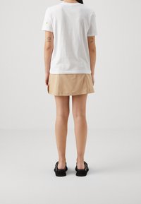 Bílá bavlněná tričko s malou výšivkou citronu na levém rukávu, kombinované s béžovou plisovanou sukní a černými botami.