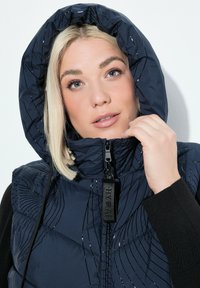 Blond kvinna som bär en marinblå huvrad quiltad jacka, håller dragkedjan nära hakan, med en neutral vit bakgrund.