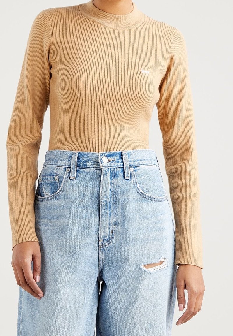 Femme portant un haut beige à manches longues côtelé rentré dans un jean bleu clair déchiré taille haute avec une déchirure sur la cuisse droite.