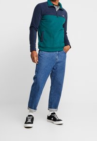 Felpa con cappuccio a blocchi di colore blu navy e verde petrolio, con collo a zip e tasche laterali. Indossata con jeans azzurri a taglio comodo e sneaker nere.