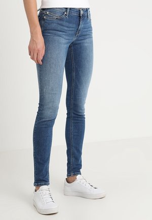 Slim fit blauwe jeans gedragen met witte sneakers en een wit topje, getoond van taille tot voeten tegen een effen achtergrond.
