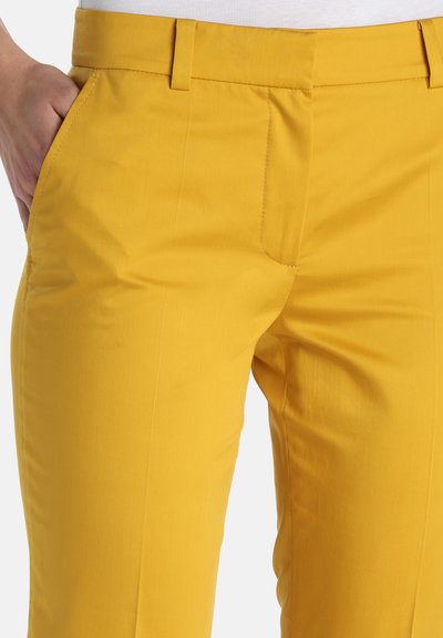 Pantalon jaune vif avec passants de ceinture et poches latérales, porté avec un haut blanc, main posée de manière décontractée dans la poche gauche.