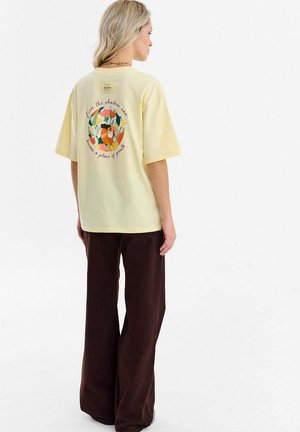 Żółta bawełniana koszulka z krótkim rękawem, z kolorową, kwiatową grafiką na plecach i tekstem wokół niej. W połączeniu z brązowymi, szerokimi spodniami.