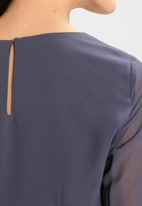 Žena v tmavě šedé blůze s malým vertikálním otvorem na zadním výstřihu, který odhaluje kousek pleti.