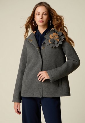 Woman wearing a gray coat with large flower decoration on shoulder, gold hoop earrings, dark pants, and hand resting near coat buttons.