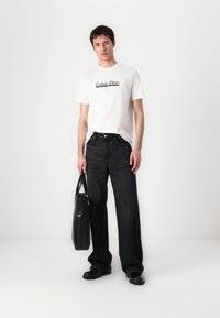 Camiseta de algodón blanca con logo negro de "Calvin Klein", combinada con jeans de mezclilla negros y holgados. El modelo sostiene un bolso tote de cuero negro y lleva zapatos negros.