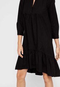 Musta mekko, jossa on V-pääntie, kolmivartaloiset hihat ja kerrostettu röyhelöhelma. Valmistettu pehmeästä kankaasta, rentoa istuvuutta.