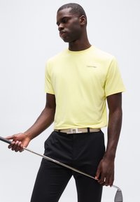 Άνδρας με κίτρινο πουκάμισο Calvin Klein και μαύρο παντελόνι κρατάει μπαστούνι γκολφ, κοιτάζοντας προς τα κάτω, στέκεται μπροστά σε απλό γκρίζο φόντο.