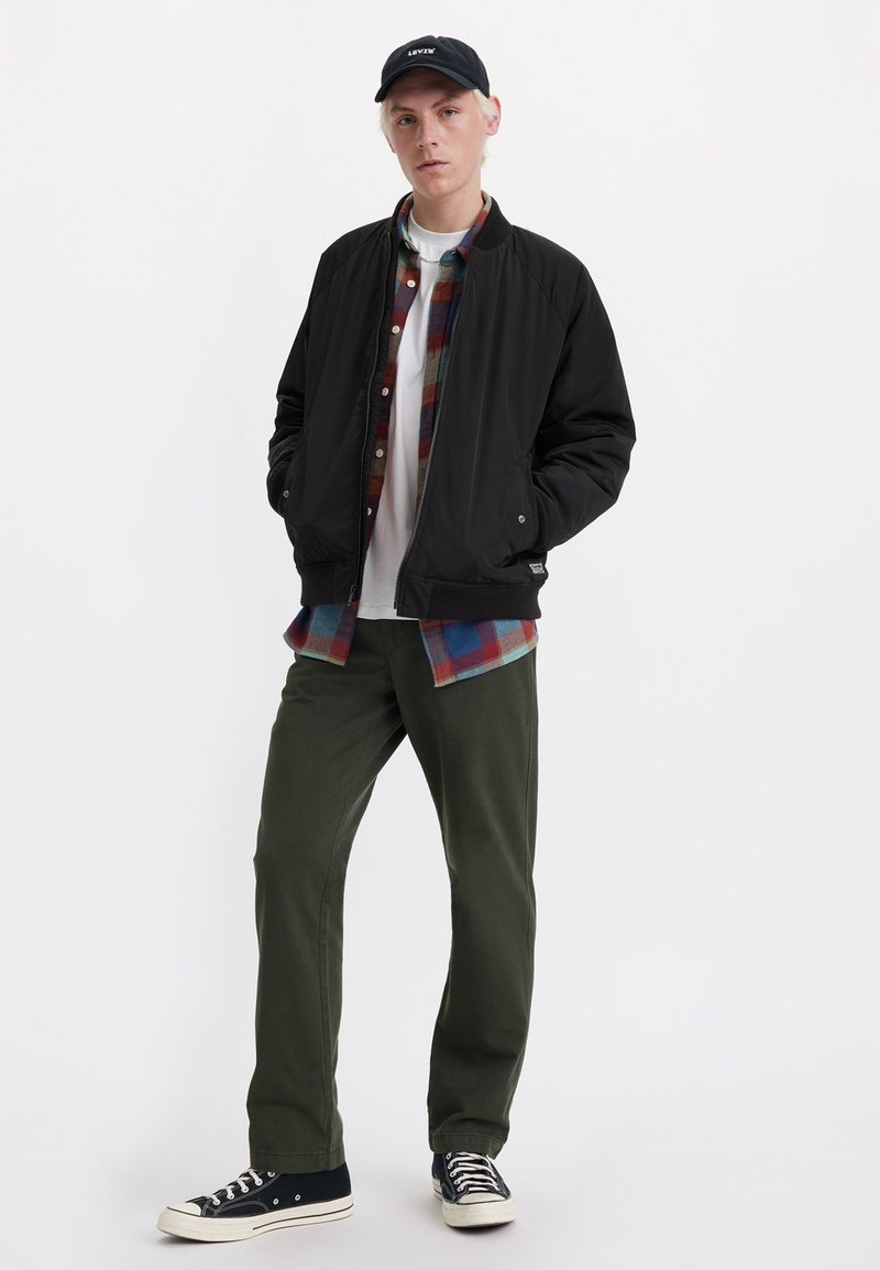 Chaqueta bomber negra con cremallera, sobre una camisa a cuadros en rojo, azul y verde. Pantalones verde oliva y zapatillas negras completan el conjunto.