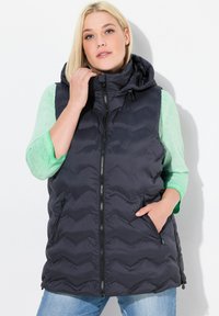 Dunkelgraue Steppweste mit Kapuze, mit horizontalem Steppmuster, vorderem Reißverschluss und zwei Seitentaschen. Über einem grünen Langarmoberteil getragen.