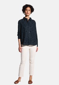 Camisa azul marino de botones y bordados con manga larga, con diseños recortados; combinada con pantalones color crema y sandalias marrones.