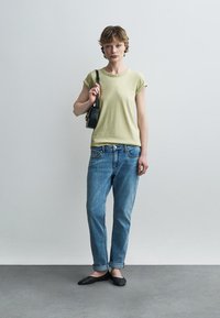 Fiatal személy világoszöld pólóban, felhajtott kék farmerban, fekete lapos cipőben, kezében egy kis fekete válltáskával, egyszerű háttér előtt.