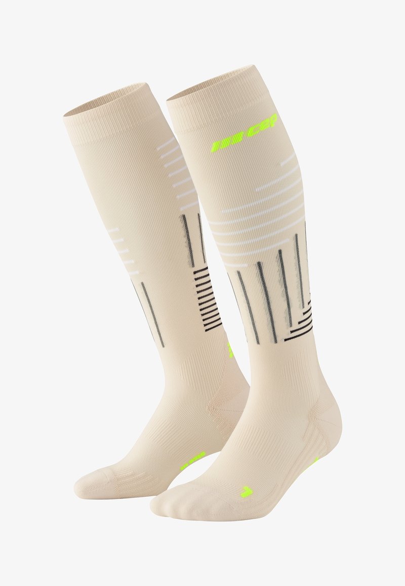 Beige compressiekousen van een gestructureerde stof. Voorzien van zwart-witte strepen en neon groene accenten op de tenen en logo's.