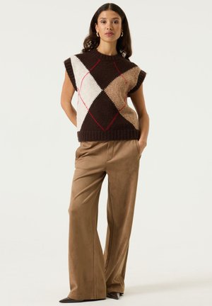 Woman stands with hands in pockets wearing wide-leg tan trousers and a sleeveless, brown, beige, and white argyle sweater vest.