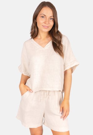 Femme aux longs cheveux bruns portant un haut beige ample à col en V et un short assorti, une main dans la poche, sur fond blanc.