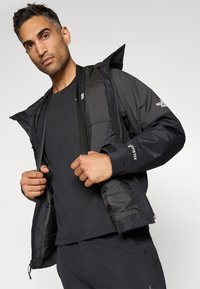 Muž upravující otevřenou černou bundu Gore-Tex North Face přes černé tričko na světle šedém pozadí.