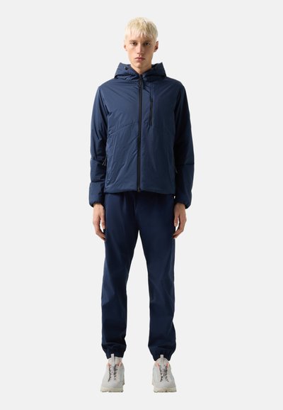 Young person with short blonde hair wearing a navy hooded jacket, matching pants, and white sneakers, standing against a plain background.