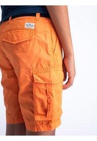Oranje cargoshorts met meerdere zakken, een label op de tailleband en een opgerolde zoom. De stof lijkt lichtgewicht en lichtjes textuur te hebben.