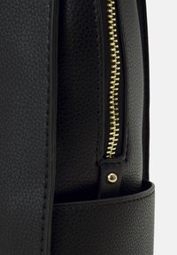 Černá taška z texturované kůže s pozdobeným zlatým zipem, zaobleným tvarem a kruhovým detailem na boku. Detailní záběr na zip a zdobení.