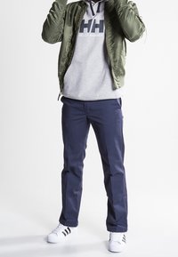Giacca bomber verde, felpa grigia con logo, pantaloni blu scuro e sneakers bianche con strisce nere. Outfit casual con un look a strati.