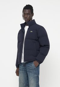 Tamno plava puf jakna s visokim ovratnikom i zatvaranjem na zip, koja ima mali zeleni Lacoste logo na prsima. Glatka, quilted tekstura.