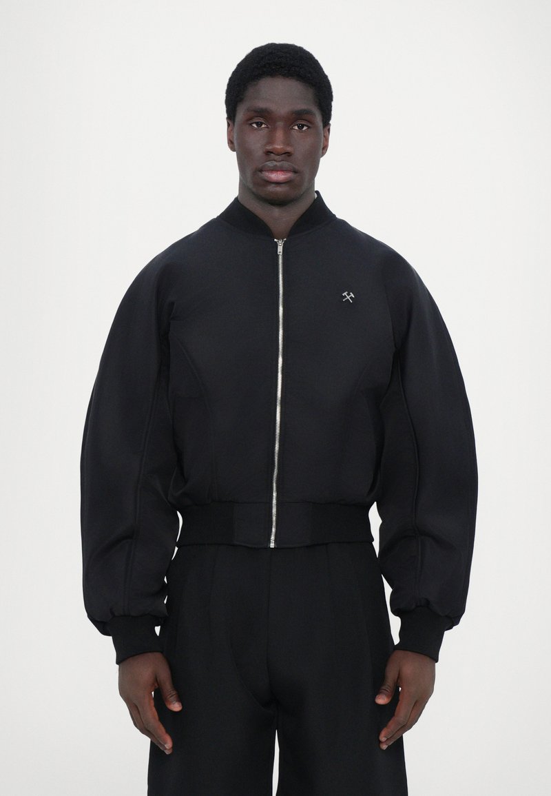 Veste bomber noire avec fermeture éclair à l'avant, poignets côtelés et coupe ajustée. Fabriquée à partir d'un tissu lisse avec un petit logo sur la poitrine.
