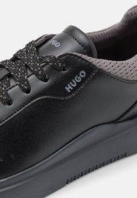 Svart lädersneaker med texturerade grå mesh-accenter, rund tå och platta svarta snören. Har en präglad logotyp på sidan.