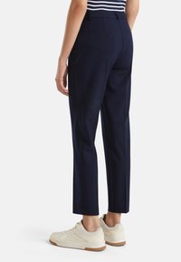 Navy úzce střižené kalhoty s plochým předním dílem, bočními kapsami a hladkým textilním povrchem. Nosit s bílými teniskami.